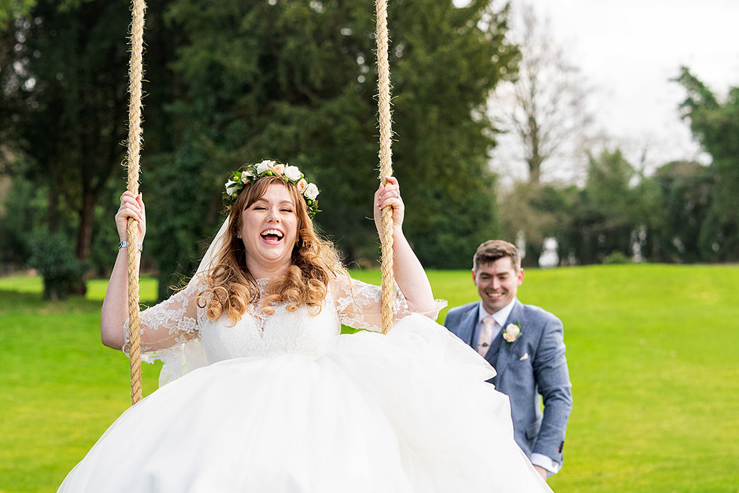 A Bride sits laughing high up on a big swing with the groom pushing her from behind