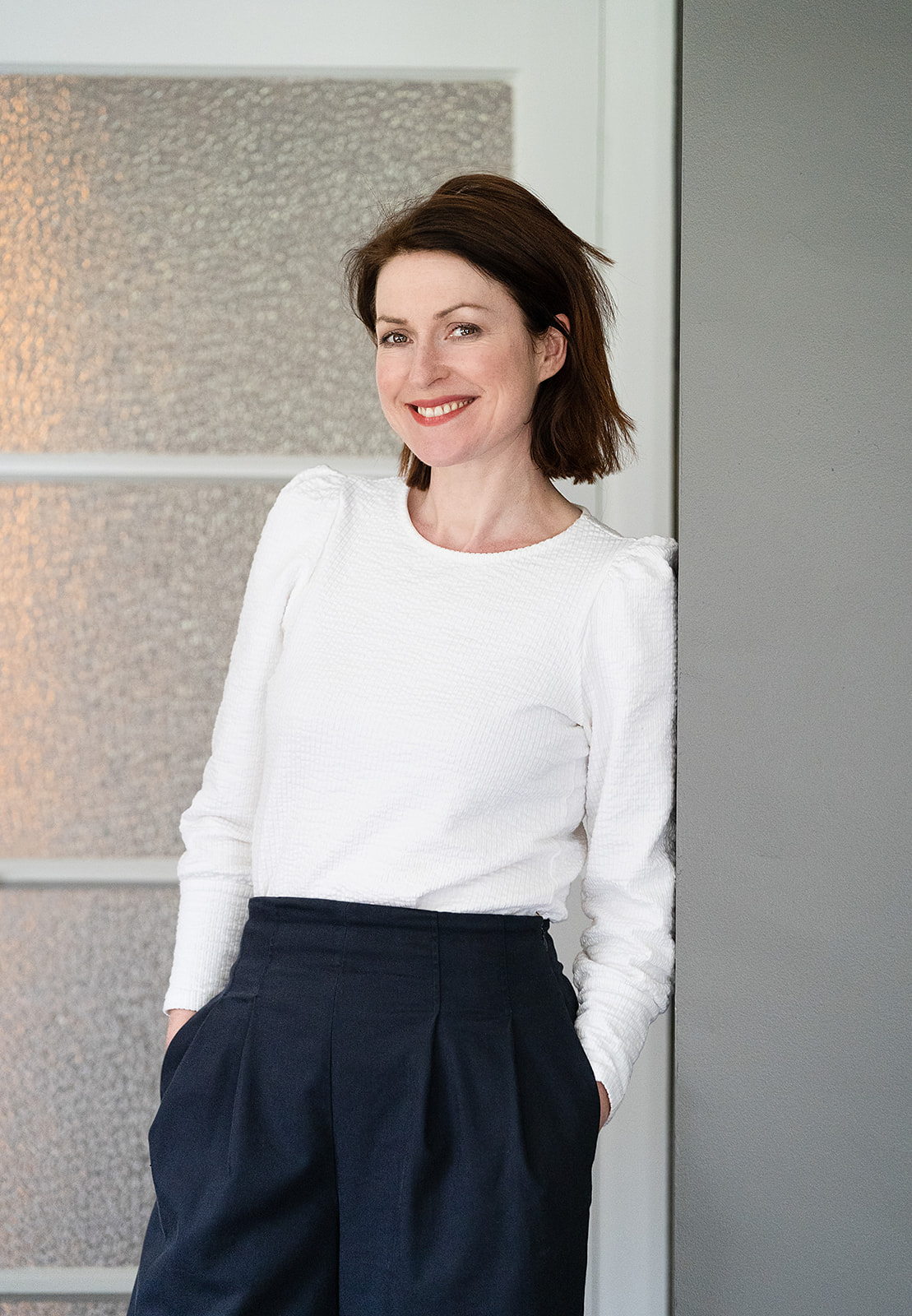 a lady dressed in white leans against a door frame and smile at the camera